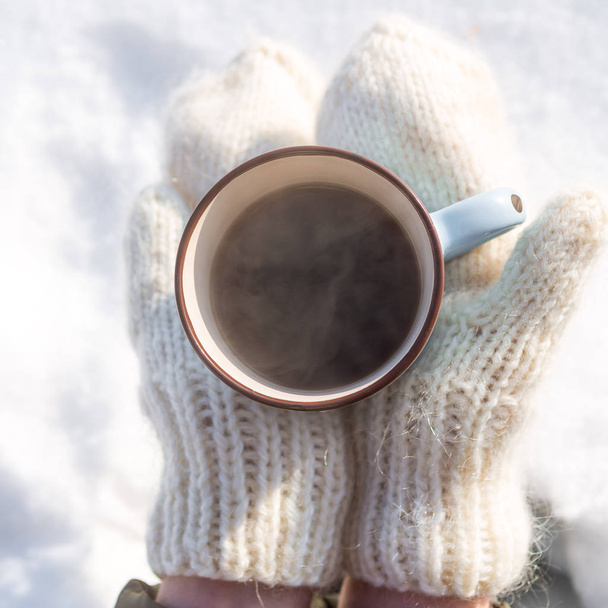 Fashion beautiful woman in hipster white knitted mittens drink hot tea on a bright winter day. Close-up of a cup with hot liquid in hand. Trees in pure first snow on a background. Top view. - Foto, afbeelding