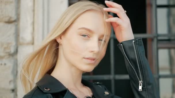 fashion portrait of a beautiful young woman with blue eyes and blonde hair in a leather jacket outdoors - Filmati, video