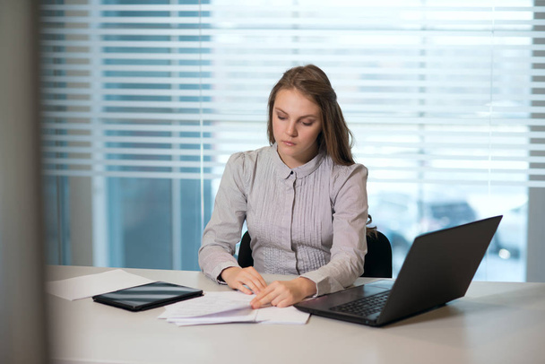 молодая девушка-предприниматель в повседневной одежде, сидящая за столом внимательно смотрит на документы, работает за компьютером
 - Фото, изображение
