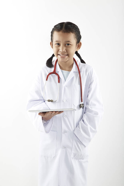 Симпатичная азиатская маленькая девочка в костюме доктора со стетоскопом и планшетом улыбается и смотрит в сторону на белом фоне, концепция здравоохранения
 - Фото, изображение