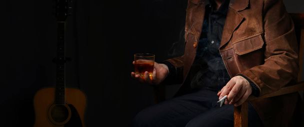 Panorama z postacią muzykiem kurtka z zamszu z papierosa i szklanką whisky na tle gitara akustyczna - Zdjęcie, obraz