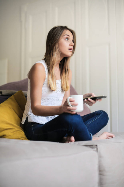 Young woman sitting on a sofa with a cup of drink and a smartphone. - Photo, Image