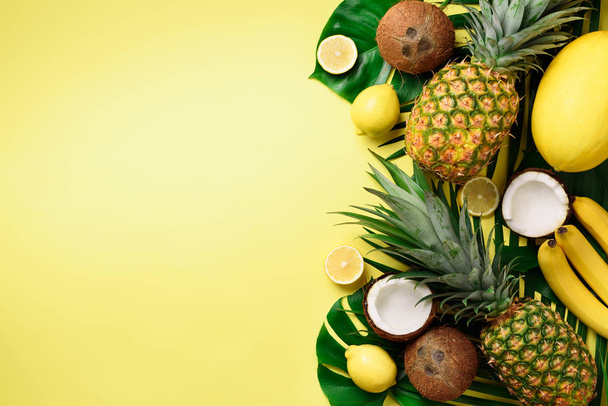 Exotic pineapples, ripe coconuts, banana, melon, lemon, tropical palm and green monstera leaves on yellow background with copyspace. Creative layout. Monochrome summer concept. Flat lay, top view. - Photo, image