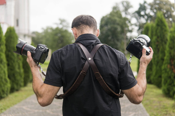 Professional wedding photographer in black shirt and with shoulder straps holding cameras like a guns against the green garden. Wedding photography. - Φωτογραφία, εικόνα