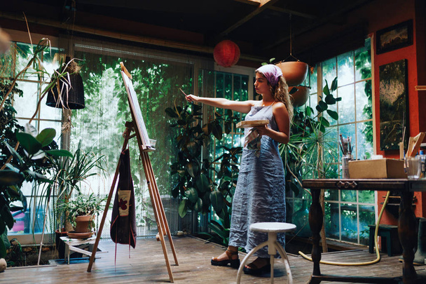 Εσωτερική βολή του επαγγελματία θηλυκό καλλιτέχνη ζωγραφικής σε καμβά στο studio με φυτά. - Φωτογραφία, εικόνα