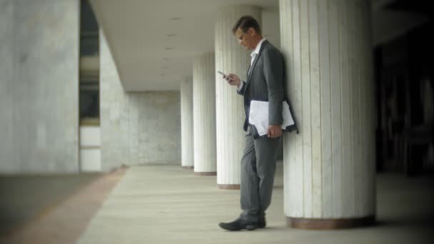 Businessman in the city. He wears a suit and briefcase. He looks through documents and talks on the smartphone - Imágenes, Vídeo