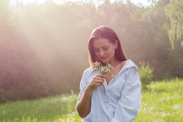 Beyaz gömlekli güzel genç bir kadın yaz mevsiminde ormanın arka planındaki bir çayırda güneş ışığında yabani çiçekler tutuyor.. - Fotoğraf, Görsel