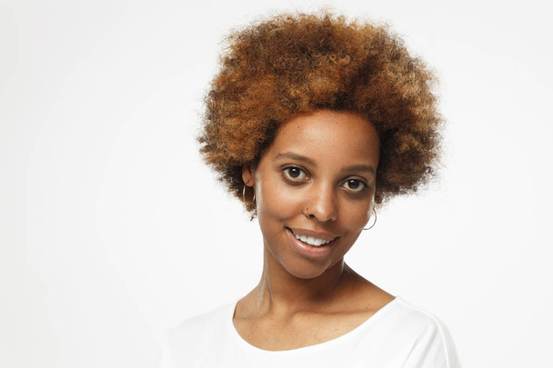 Closeup photo of young african american woman isolated on gray background with copyspace, dressed in white t shirt, turned to camera with one shoulder, smiling cheerfully - Foto, Imagen