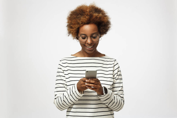 Young african american hipster millennial woman in trendy round eyeglasses, isolated on gray background, texting or browsing with happy relaxed smile - Photo, Image