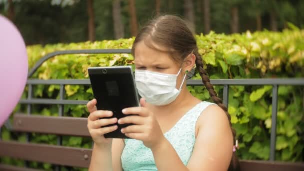 Roztomilá dívka v respirátor používá tabletu v parku na lavičce - Záběry, video