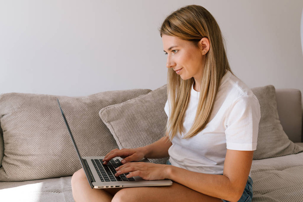 Blonde fille en vêtements décontractés en utilisant un ordinateur portable tout en étant assis sur le canapé à la maison
. - Photo, image