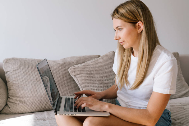 Hermosa chica en ropa casual está utilizando el ordenador portátil, y sonriendo mientras está sentado en el sofá en casa. Mujer joven en una camiseta blanca escribiendo en el teclado haciendo trabajo independiente en Internet
. - Foto, imagen