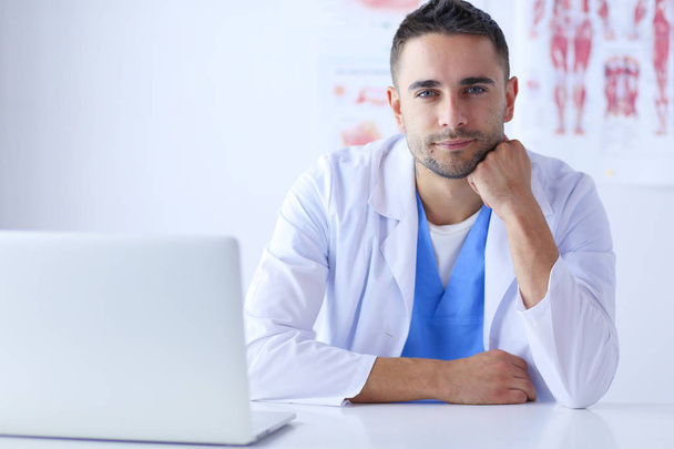 Portrait of a male doctor with laptop sitting at desk in medical office - Foto, afbeelding