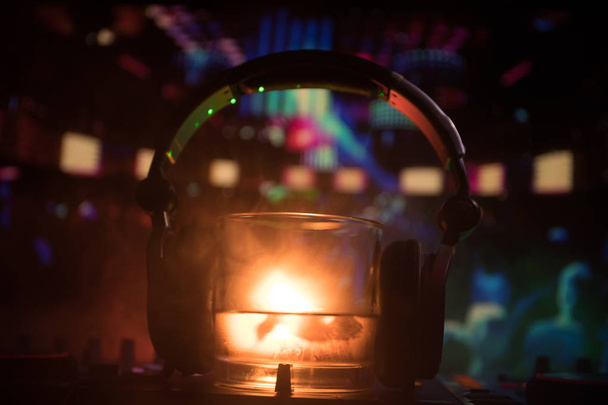 Glas met whisky met ice cube binnen op dj-controller bij nightclub. DJ koptelefoon op een glas van whilsky op muziek feest in nachtclub met disco lichten. Selectieve aandacht - Foto, afbeelding