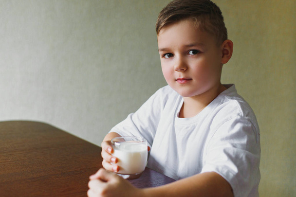 Son drinks milk in the kitchen in the morning. A young boy shows a class, a glass of tasty, homemade milk. Healthy breakfast. - Photo, Image