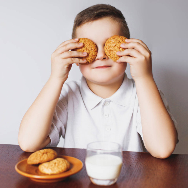 Egy fiatal fiú tartja zabliszt cookie-kat, és becsukja a szemét. A fiú ült a konyhában, a kekszek és a tej. - Fotó, kép