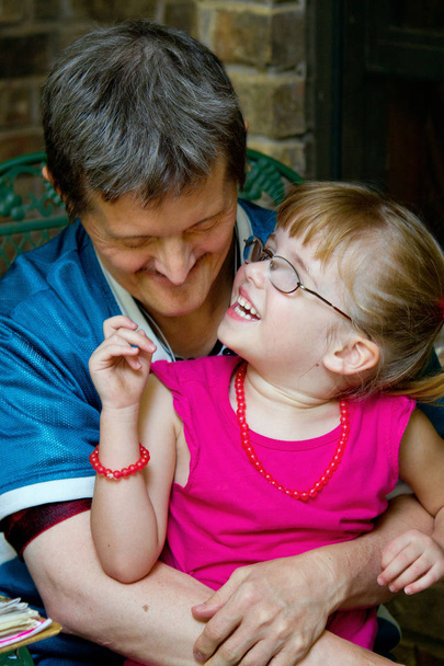 Мужчина с синдромом Дауна держит свою молодую племянницу, когда она смотрит на него, смеясь. У неё толстые очки и большая улыбка. Он смотрит на нее с улыбкой
. - Фото, изображение