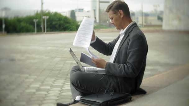 Businessman is sitting on the stairs in the city. He wears a suit and briefcase. He works on a laptop - Footage, Video