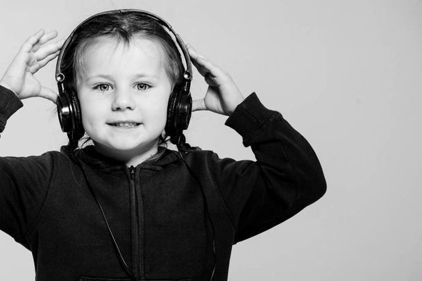 Μοντέρνο και κομψό. Ευτυχισμένο μικρό παιδί που απολαμβάνει μοντέρνα μουσική σε γκρι φόντο. Μικρό κορίτσι με κομψά ακουστικά που ακούει μοντέρνα τραγούδια. Χρήση σύγχρονης τεχνολογίας για ευχαρίστηση και μελέτη. - Φωτογραφία, εικόνα