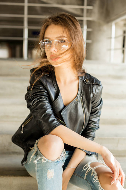 Lifestyle photo of young cheerful woman in fashion transparent glasses wearing a rock black style leather jacket look to the camera. Hipster urban style. Lifestyle outdoor portrait. - Фото, изображение