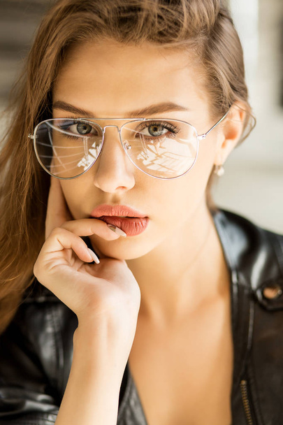 Close up lifestyle portrait of young cheerful woman in fashion transparent glasses wearing a black rock style leather jacket touch her big sexy lips. Hipster urban style. Lifestyle outdoor portrait. - Photo, image