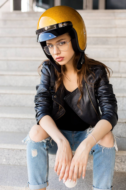 Beautiful young woman in fashion glasses, rock black style leather jacket and glitter gold motorbike helmet sit on stairs and look to camera. Hipster urban style girl. Lifestyle outdoor city portrait. - Photo, image