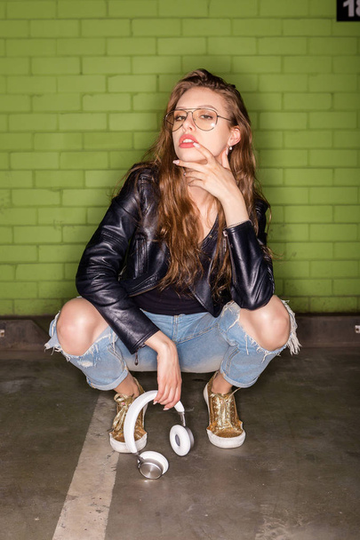 Fashion portrait of beautiful woman DJ in cool glasses, rock black leather jacket slav squat sit and with white music headphones sound. Hipster urban style girl. Lifestyle outdoor city portrait. - Foto, Imagen