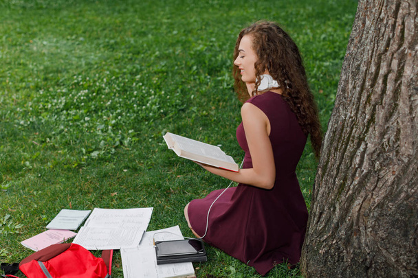 curly young beautiful girl student holding book and sitting under tree in summer park. - Foto, Imagen