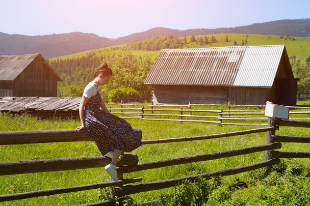 Young woman sitting on a wooden fence. Houses and green mountains in the background - 写真・画像