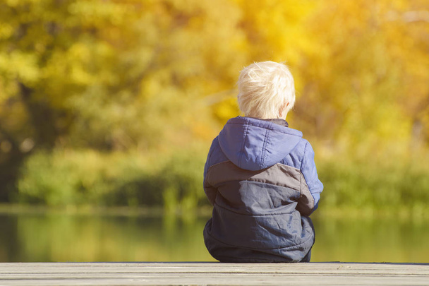 Ξανθό αγόρι κάθεται στην προβλήτα στον ποταμό. Κίτρινα φύλλα στα χέρια. Πίσω όψη - Φωτογραφία, εικόνα