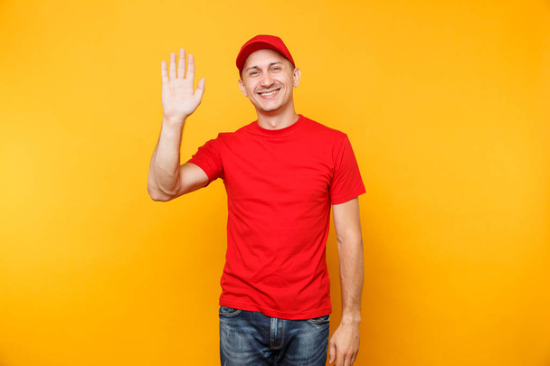 Zusteller in roter Uniform isoliert auf gelbem Hintergrund. Ein lächelnder männlicher Angestellter mit Schirmmütze, ein T-Shirt-Kurierhändler winkt und grüßt mit der Hand, als jemand eine Geste bemerkt. Dienstleistungskonzept. Kopierraum - Foto, Bild
