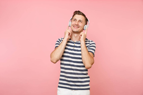Retrato de un joven sonriente con una camiseta a rayas escuchando música con auriculares inalámbricos blancos, disfrute aislado sobre un fondo rosa pastel de tendencia. Gente emociones sinceras concepto de estilo de vida
 - Foto, Imagen