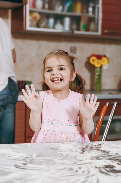 Pieni naurava lapsi tyttö vaaleanpunainen mekko leikkii, tahraa kädet jauhoja ja osoittaa kämmenet kevyt keittiö pöydässä. Lapsen tytär ruoanlaitto, evästeet viikonloppuna aamulla kotona. Hyvää lapsuutta.
 - Valokuva, kuva