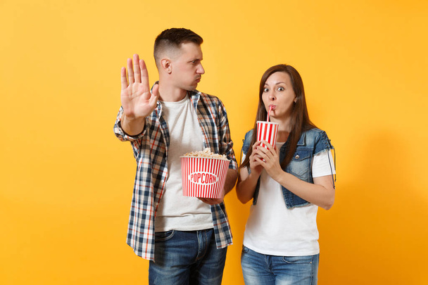 Young irritated couple woman man watching movie film on date holding bucket of popcorn and plastic cup of soda or cola showing stop gesture with palm isolated on yellow background. Emotions in cinema - Foto, afbeelding