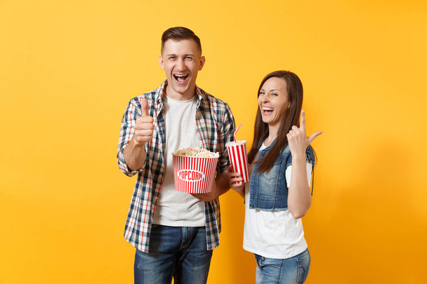 Jong paar vrouw man kijken film film lachen op datum houden emmer popcorn kunststof kopje soda cola tonen duim omhoog wijzende vinger van de index van geïsoleerde op gele achtergrond. Emoties in bioscoop - Foto, afbeelding