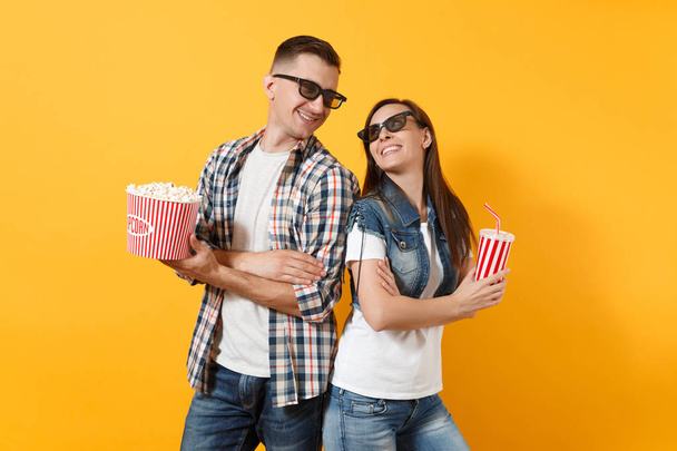 Молодая улыбающаяся пара мужчина мужчина в 3D очки смотреть фильм на свидании держа ведро попкорна пластиковой чашки соды или колы глядя друг на друга изолированы на желтом фоне. Эмоции в кино
 - Фото, изображение