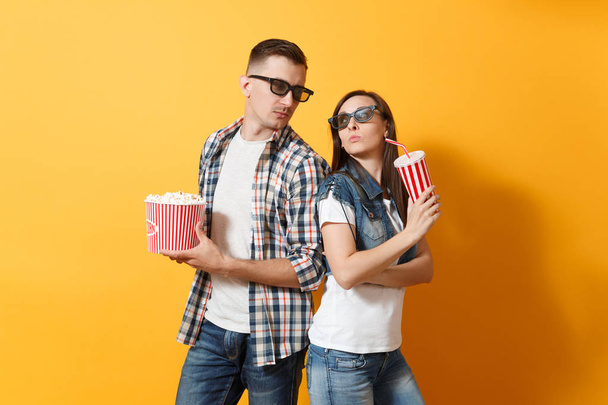 ヤングでは、ソーダやコーラが黄色の背景上に分離されて互いを見てポップコーン プラスチック カップのバケツを開催日に映画を見て 3 d メガネのカップル女性男性を懸念しています。映画の感情 - 写真・画像