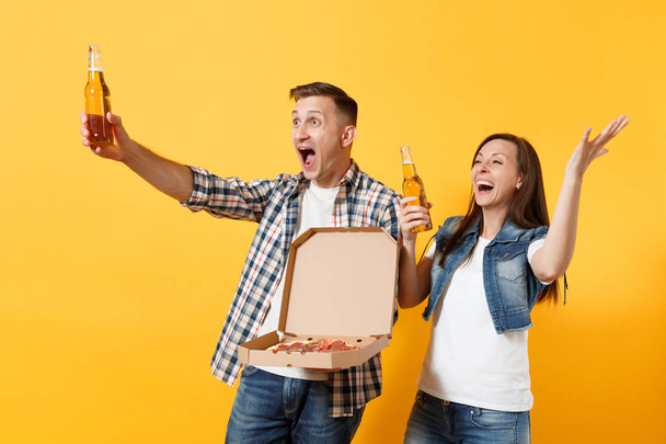 Joven alegre pareja mujer hombre deporte fans animar a equipo de apoyo sosteniendo botella de cerveza pizza italiana en caja plana de cartón extendiendo las manos aisladas sobre fondo amarillo. Deporte familia ocio estilo de vida
 - Foto, Imagen
