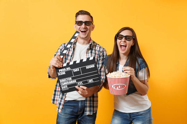 Молодая счастливая пара женщина мужчина в 3D очки смотреть фильм на свидании, держа классический черный фильм, делая доски и ведро попкорна изолированы на желтом фоне. Эмоции в концепции кино
 - Фото, изображение