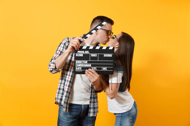 Молодая счастливая красивая пара женщина мужчина в 3D очки смотреть фильм на свидании, держа классический черный фильм, делая доски и поцелуи изолированы на желтом фоне. Эмоции в концепции кино
 - Фото, изображение