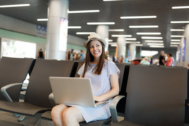 Young charming traveler tourist woman in hat working on laptop while waiting in lobby hall at international airport. Passenger traveling abroad on weekends getaway. Air travel, flight journey concept - Photo, Image