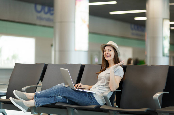 Eine junge, lachende Touristin sitzt am Laptop und breitet die Hände aus, während sie in der Lobby des internationalen Flughafens wartet. Reisende, die am Wochenende ins Ausland reisen. Flugkonzept - Foto, Bild