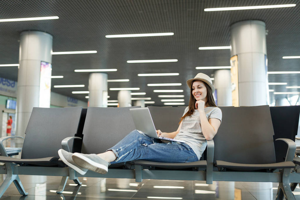 Νεαρή γοητευτική γυναίκα τουριστικά ταξιδιώτη στο καπέλο που εργάζονται στον φορητό υπολογιστή ενώ περιμένετε στο λόμπι hall στο διεθνές αεροδρόμιο. Επιβάτες που ταξιδεύουν στο εξωτερικό για απόδραση το Σαββατοκύριακο. Αεροπορικά ταξίδια, πτήση ταξίδι έννοια - Φωτογραφία, εικόνα