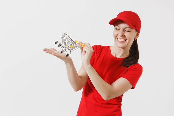 Delivery woman in red uniform isolated on white background. Female courier or dealer in cap, t-shirt, jeans holding supermarket grocery push cart for shopping on palm. Copy space for advertisement - Photo, Image