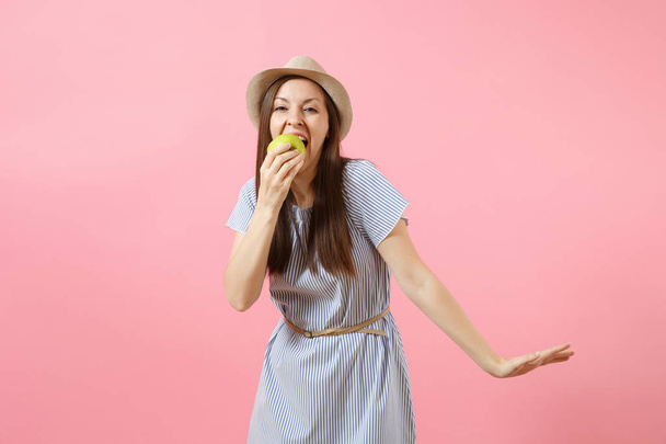 青いドレスの若い美しい女性の肖像画の夏麦わら帽子持株、ピンクの背景に分離された緑の新鮮なリンゴの果実を食べるします。健康的なライフ スタイル、人々、心から感情の概念。コピー スペース - 写真・画像
