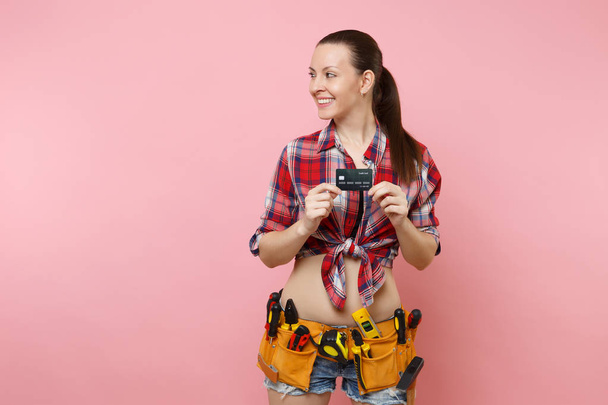 Mladá usměvavá žena údržbář v kostkované košile, džínové šortky, nástroje kit pás plný různých nástrojů drží bankovní kreditní kartu izolované na růžovém pozadí. Žena dělá mužské práce. Koncept renovace - Fotografie, Obrázek