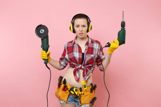 手袋でエネルギーの便利屋女性, ノイズ絶縁ヘッドフォン、保持力の楽器の完全キット ツール ベルト見たピンクの背景に分離された電気ドリル。男性の仕事の女性。改修工事コンセプト - 写真・画像
