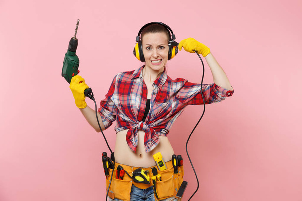 手袋でエネルギー セクシーな便利屋女性, ノイズ絶縁ヘッドフォン、キット ツール ベルト ピンクの背景に分離電源電気ドリルの保持器の完全な。男性の仕事の女性。改修工事コンセプト - 写真・画像