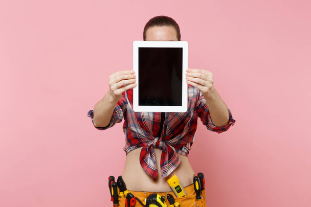 Женщина с комплектом инструментов пояс полный разнообразных инструментов держать планшетный компьютер с пустым черным пустым экраном сенсорный экран изолирован на розовом фоне. Женщина занимается мужской работой. Концепция реконструкции
 - Фото, изображение