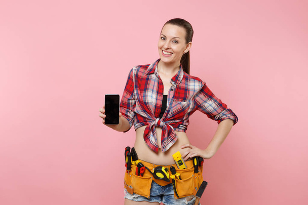 Woman with kit tools belt full of instruments holding showing camera mobile phone with blank black empty screen display touchscreen isolated on pink background. Female male work. Renovation concept - Foto, Imagen